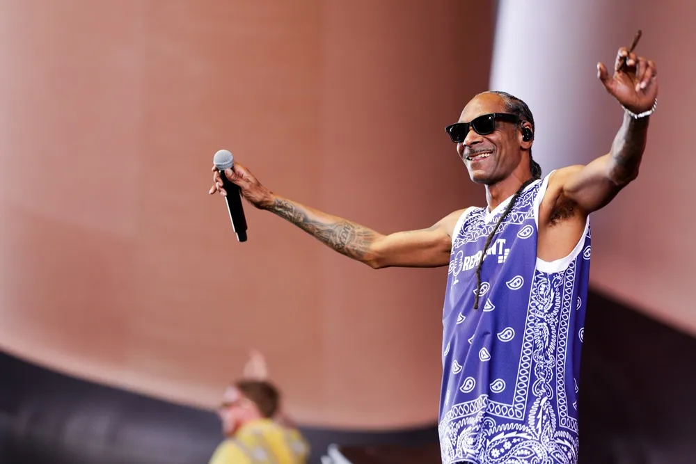 Sahnedeki neşeli Snoop Dogg, sol elinde mikrofon tutarken sağ kolunu havaya kaldırmış, elinde ince sarılı bir nesneyle, mavi beyaz bandana desenli bir atlet ve güneş gözlüğüyle geniş bir gülümsemeyle Coachella performansını sergiliyor.
