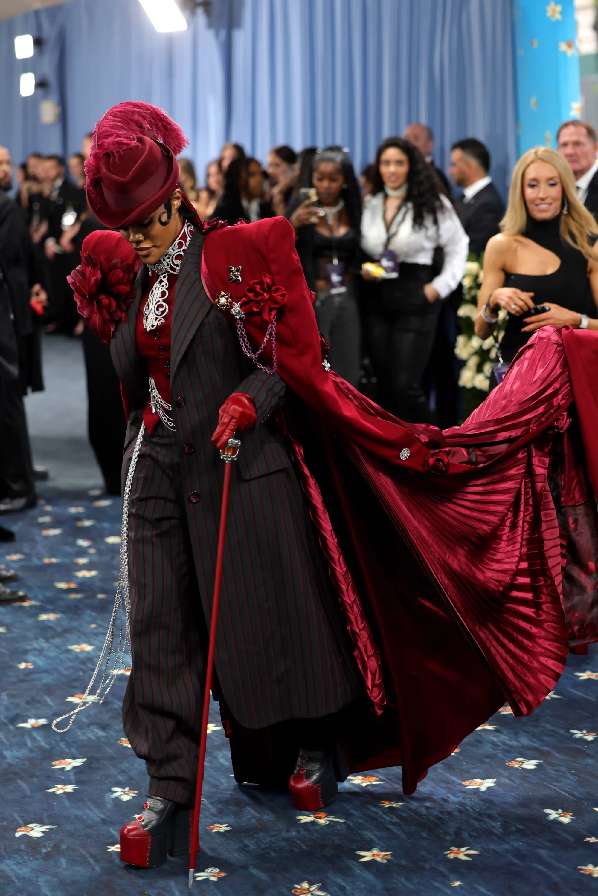 Kırmızı tüylü geniş kenarlı şapkası, siyah üzerine ince kırmızı çizgili özel dikim takım elbisesi ve göz alıcı, pileli kırmızı peleriniyle Met Gala'da boy gösteren Rihanna, zarif bastonu ve parlak takılarıyla dikkat çeken, kişiselleştirilmiş bir stil sunuyor.