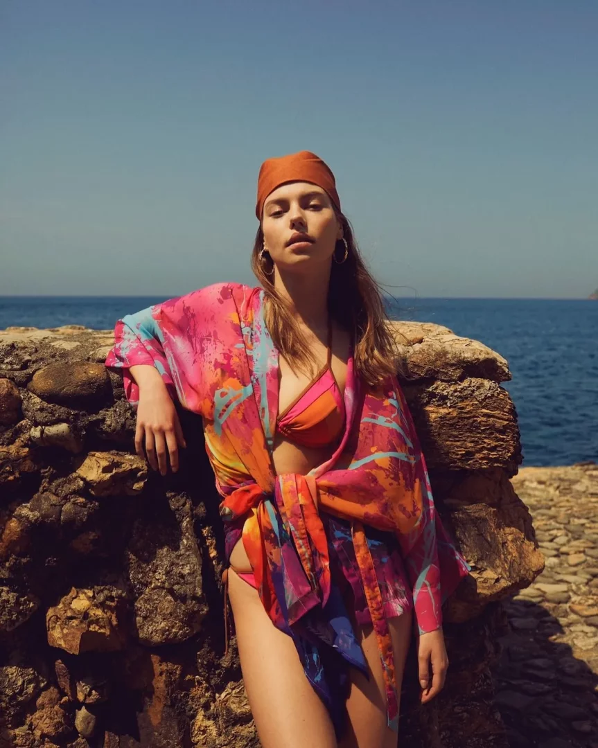 Mavi gökyüzü ve denizin önünde, kayalık bir duvara yaslanmış, turuncu bandana ve altın halka küpeler takan bir model, Minca markasına ait pembe, turuncu, mavi ve mor tonlarında soyut desenli bir bikini ve bağlamalı bir kimono giyiyor.
