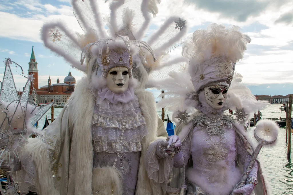 Venedik Karnavalı’nda maskeli ve gösterişli beyaz kostümler giymiş iki katılımcı – San Giorgio Maggiore manzarası