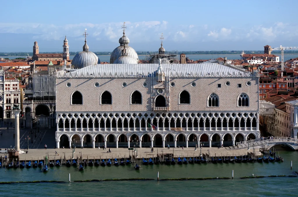 Venedik'in ikonik yapılarından Dükler Sarayı (Palazzo Ducale), su kenarında Gotik detaylarla bezeli açık renk cephesiyle tüm ihtişamıyla yükselirken, önündeki rıhtımda geleneksel gondolların sıralandığı, masmavi gökyüzü altındaki büyüleyici bir panorama.