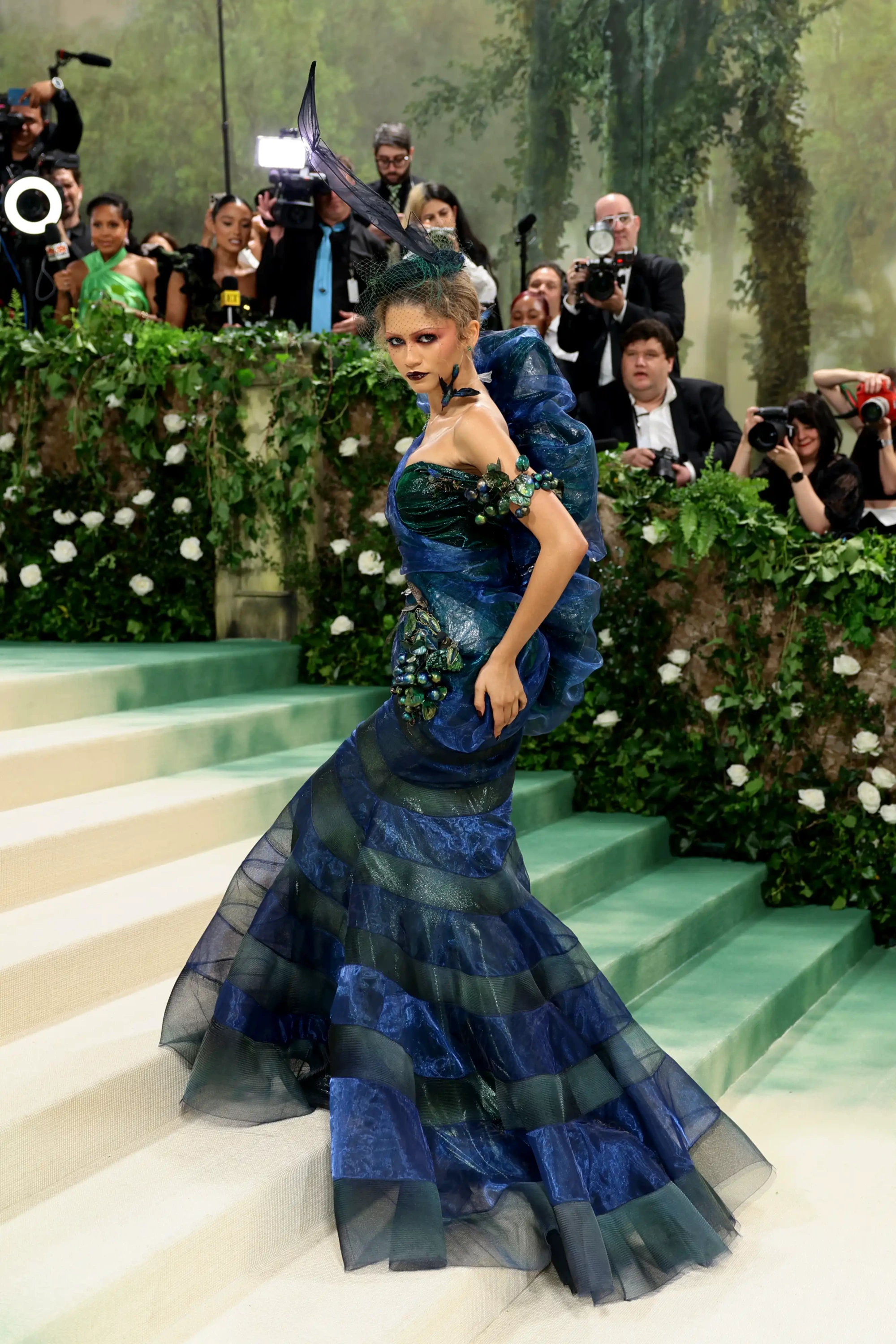 Zendaya, Met Gala 2024'te Zamanın Bahçesi temasını yansıtan, mavi ve yeşil tonlarda şeffaf katmanlı kumaş detayları, kelebek kanadına benzer bir başlık ve vücudunda böcek/bitki motifleriyle süslenmiş çarpıcı elbisesiyle, yeşilliklerle çevrili merdivenlerde poz veriyor.