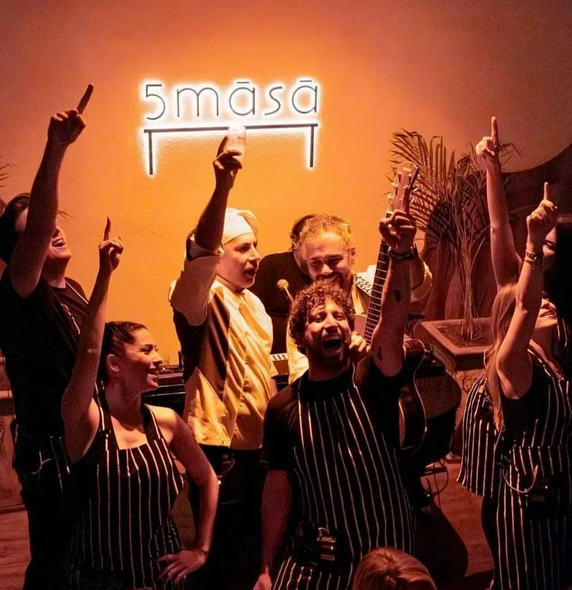 5 masa restoran ekibi, canlı müzik eşliğinde neşeli bir anda, şef önlüğü ve çizgili önlükleriyle, duvardaki neon ışıklı 5māsā tabelasının önünde ellerini havaya kaldırarak coşkuyla tezahürat yapıyor, bir yandan da gülümseyerek kutluyor.