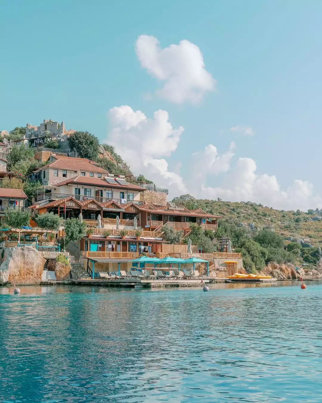 Ankh Pansiyon'un yeşil yamaçlara basamaklı bir şekilde konumlanmış, teraslı ve ahşap detaylı binaları, önünde turkuaz denizin ve şezlonglu, mavi şemsiyeli bir platformun bulunduğu pitoresk bir kıyı manzarasını sergiliyor.