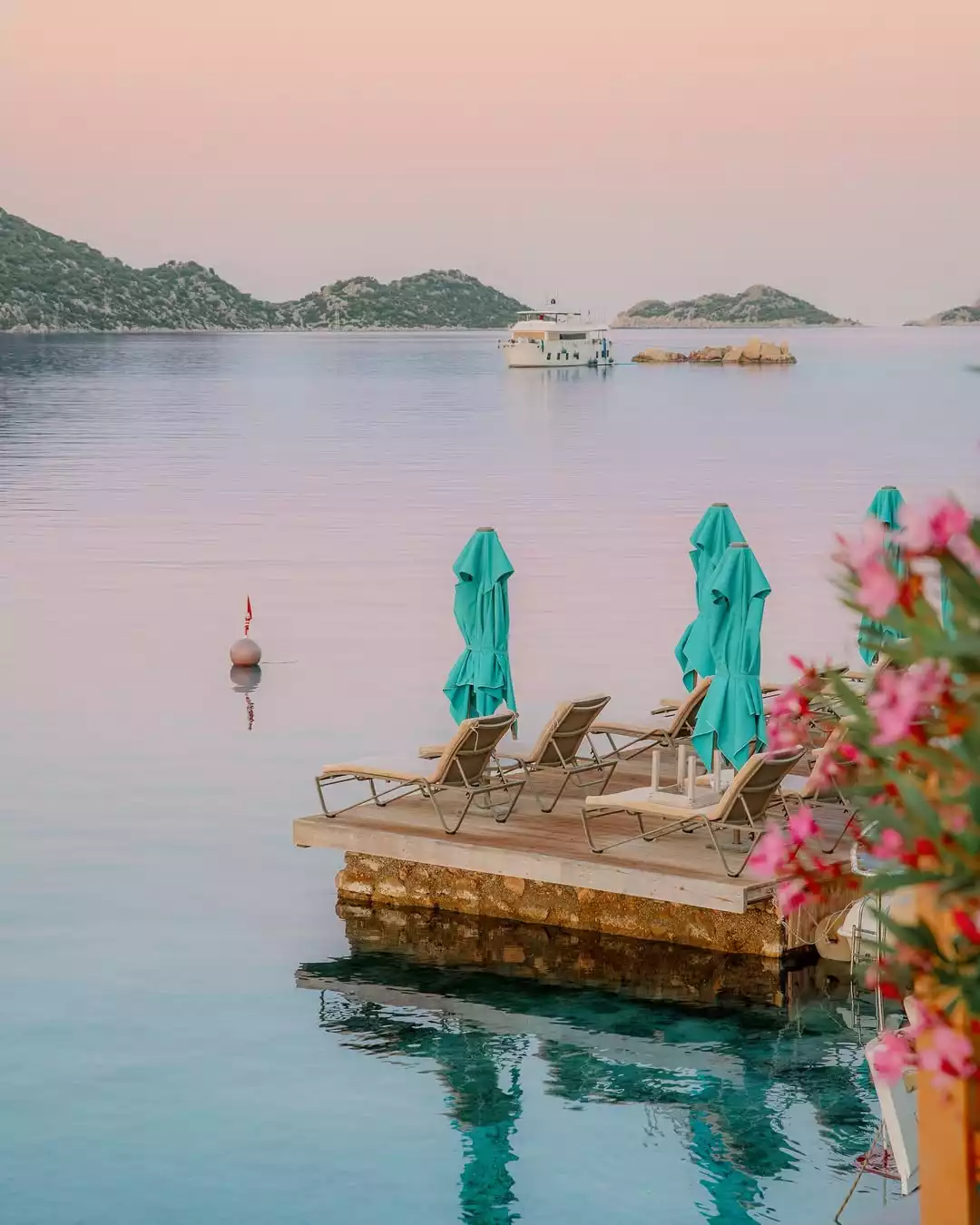 Pastel pembe bir gökyüzünün yansıdığı sakin denizde, iskelenin üzerinde turkuaz renkli katlanmış şemsiyeler ve boş şezlonglar sıralanmış, karşıda yemyeşil adacıklar arasında beyaz bir yat demirlemiş dururken, sağ ön planda odak dışı pembe çiçekler bu dingin manzarayı tamamlıyor.