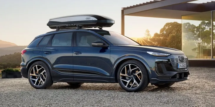 Güneşin vurduğu modern bir yapı ve uzaktaki dağlık arazi önünde çakıllı bir zeminde park etmiş, tavan kutusu takılı koyu mavi Audi Q6 e-tron elektrikli SUV, 625 km menziliyle en uzun menzilli elektrikli SUV modelleri arasında dikkat çekiyor.