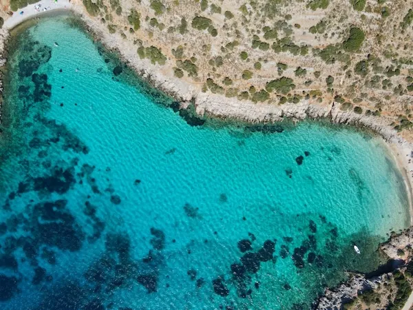 Oggusto Çocuklu Aileler İçin Yunan Adaları Rehberi: Chios kıyılarında, ailece güvenle yüzülebilecek berrak turkuaz sulara sahip korunaklı sakin bir koy. Sığ denizin altındaki koyu kayalıklar yukarıdan belirgin şekilde izlenirken, demirlemiş küçük beyaz tekne adanın huzurunu yansıtıyor.
