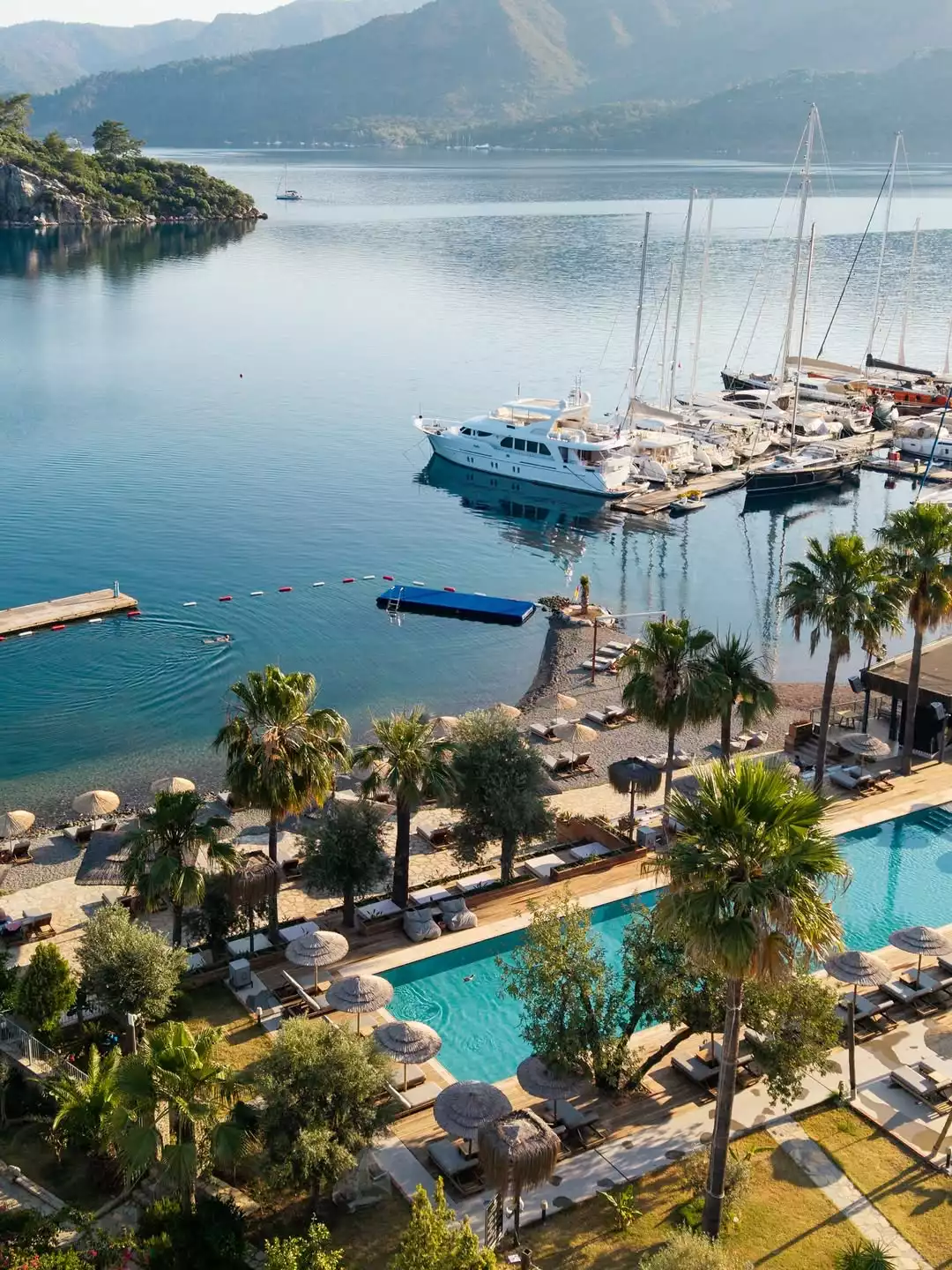 Cooks Club otelinin büyüleyici manzarasını yansıtan bu yüksek açılı fotoğrafta, berrak turkuaz denizde demirli lüks yatlar ve yelkenlilerle dolu bir marina, şemsiyeler ve şezlonglarla süslenmiş bir plajın hemen yanında uzanan, palmiye ağaçları ve yeşil bitkilerle çevrili, pırıl pırıl bir yüzme havuzu göz kamaştırıyor.