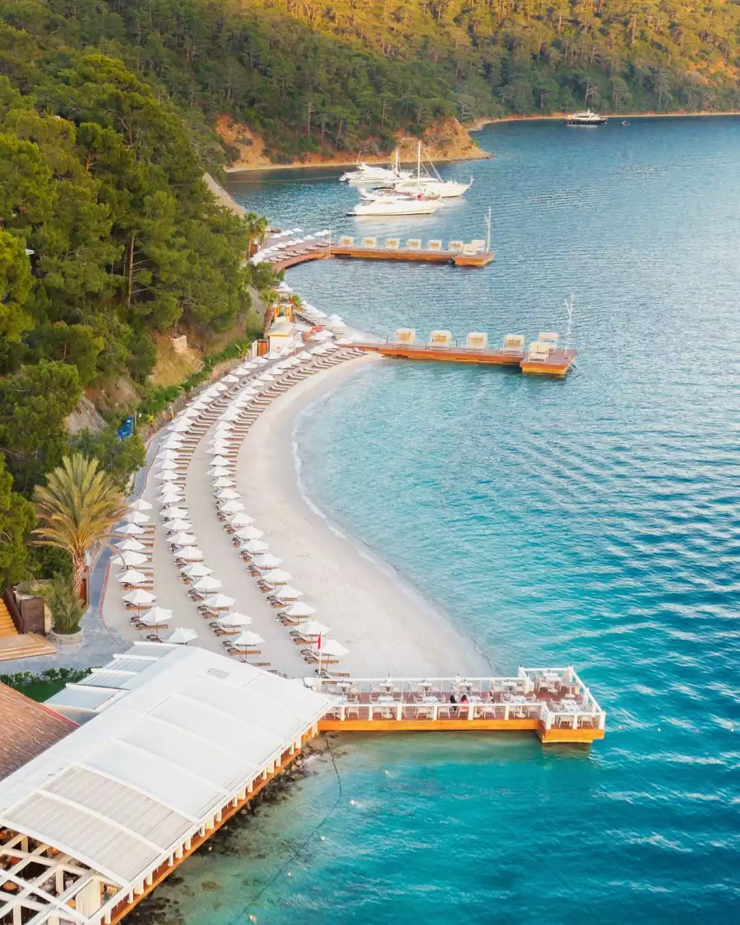 D-Resort'un, beyaz şemsiyelerle dolu uzun kumlu plajının, turkuaz sulara uzanan iskelelerin ve arkasında yemyeşil tepelerin görüldüğü kuşbakışı görüntüsü, su üzerindeki restoranlarıyla birlikte misafirlerine eşsiz bir tatil deneyimi sunuyor.