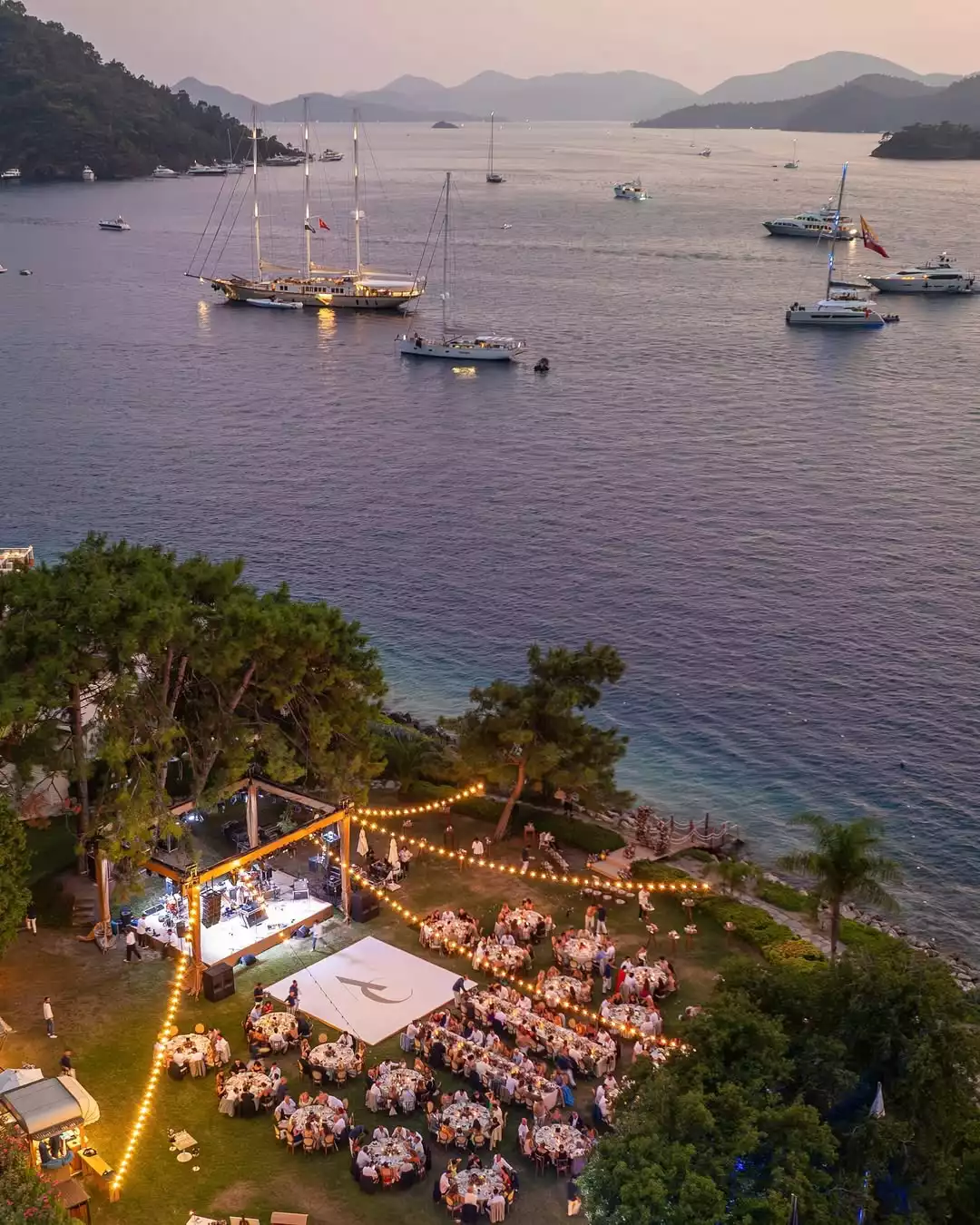 Akşam alacakaranlığında deniz kenarındaki yemyeşil bir çim alanda, parlayan ışık zincirleriyle aydınlatılmış sahne ve davetli masalarıyla dolu, uzakta birçok yatın demirli olduğu büyüleyici bir açık hava etkinliği alanı.