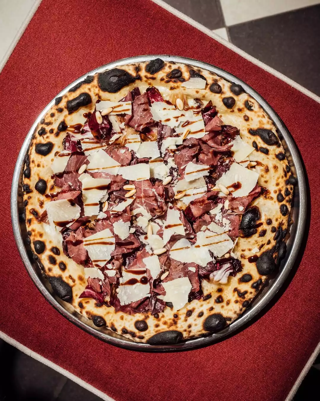 İstanbul'un en iyi pizzacıları listesinden bir sunum olan bu Napoliten tarzı odun fırını pizzası, karakteristik kömürleşmiş kenarlarıyla dikkat çekerken, üzerinde ince dilimlenmiş füme et, rendelenmiş Parmesan peyniri, hafif acımsı kırmızı marul yaprakları ve çam fıstıkları ile zenginleştirilmiş balzamik sos gezdirilmiş halde iştah açıcı bir görüntü sunuyor.
