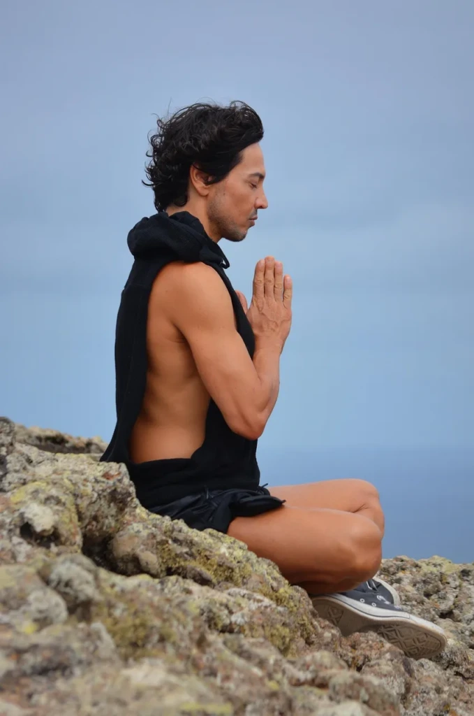 Kayalıklar üzerinde bağdaş kurarak meditasyon yapan, siyah atlet giymiş bir adam; doğayla bütünleşmiş sessiz bir farkındalık anı.