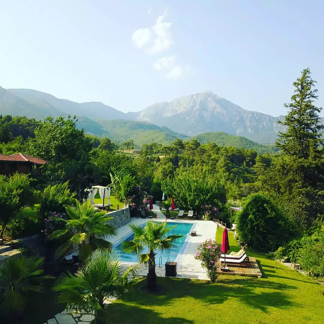 Hotel Lycia'nın yemyeşil ağaçlar ve palmiyelerle çevrili, berrak mavi suya sahip dikdörtgen yüzme havuzu, şezlonglar ve kırmızı şemsiyelerle donatılmış geniş dinlenme alanı, arka planda yükselen görkemli dağlarla birlikte huzurlu bir tatil ortamı sunuyor.