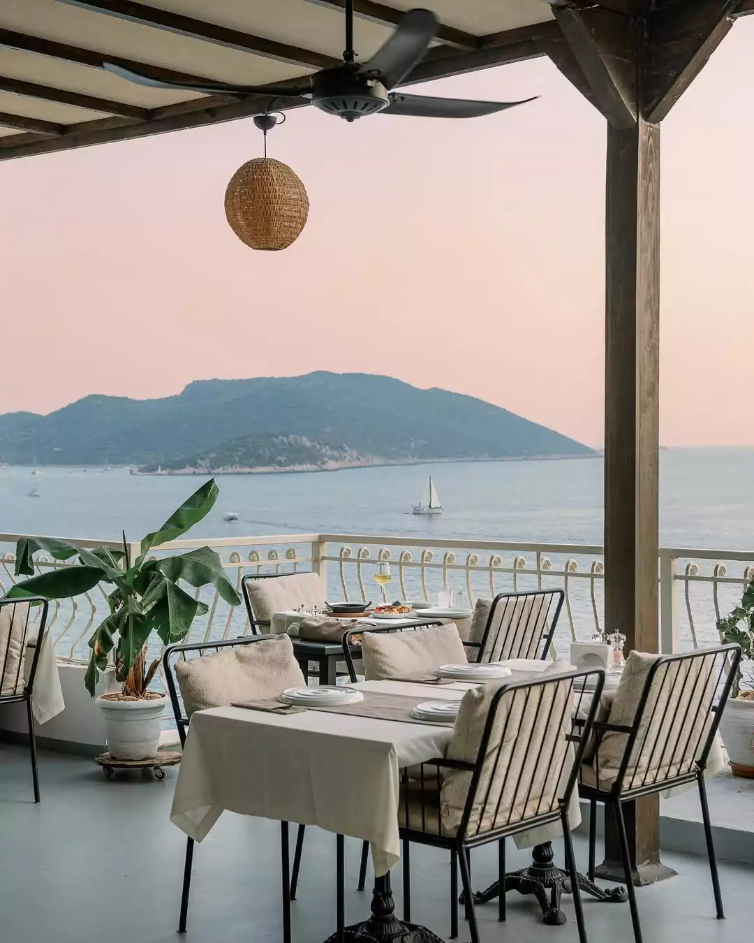 Otel terasında, gün batımının yumuşak pembe tonlarında Akdeniz'e nazır, şık masaların ve konforlu sandalyelerin hazırlandığı, uzakta bir yelkenlinin süzüldüğü ve yemyeşil adaların çevrelediği huzurlu bir yemek deneyimi sunan manzara.