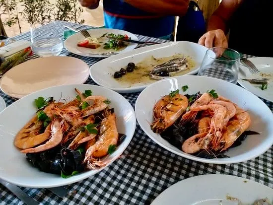 Damalı masa örtüsü serili bir yemek masasında, ana odak noktası olarak iki beyaz oval tabakta, siyah mürekkep balığı soslu makarna veya risotto üzerinde taze maydanozlarla süslenmiş bol miktarda ızgara karides sunumu dikkat çekiyor; arka planda ise kısmen yenmiş hamsi, zeytin ve küçük salata tabakları ile misafirlerin elleri, keyifli bir Akdeniz yemeği atmosferini tamamlıyor.