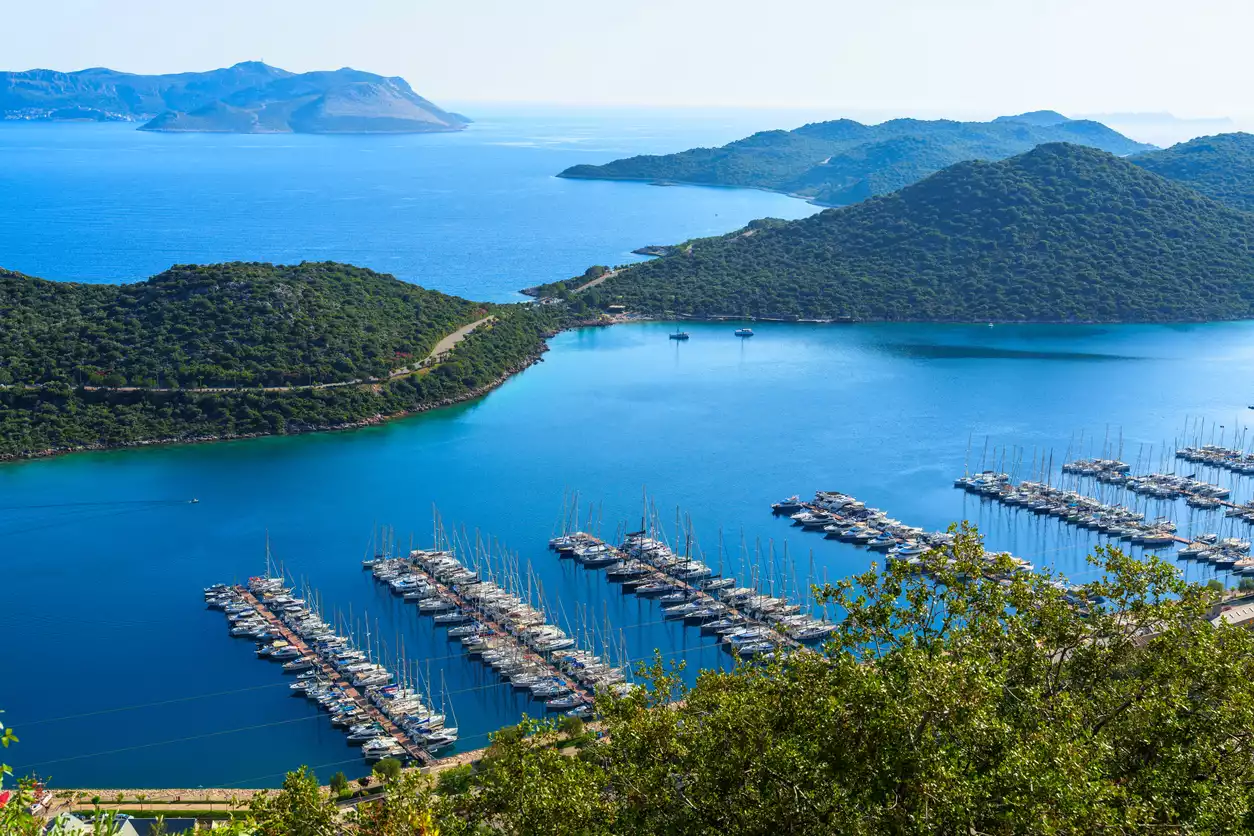 Kaş'ın sakin koylarından birinde çekilen bu geniş açılı manzarada, masmavi berrak sular üzerinde düzgün sıralar halinde bağlı çok sayıda yelkenli ve motor yat ile yemyeşil ağaçlarla kaplı kıyı şeridi ve uzaklardaki adalar net bir şekilde görünüyor.