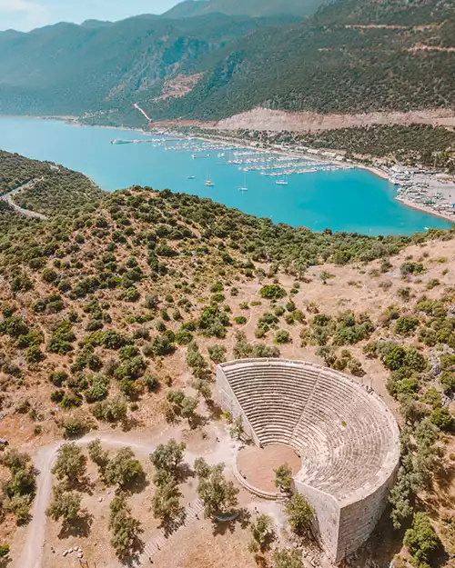 Kaş'ın eşsiz manzarasını sunan bu hava fotoğrafında, kuru otlar ve zeytin ağaçlarıyla çevrili iyi korunmuş antik tiyatro ön plandayken, Akdeniz'in masmavi sularında yüzen teknelerle dolu canlı marina ve ardındaki heybetli dağlar doğal güzellikleri sergiliyor.