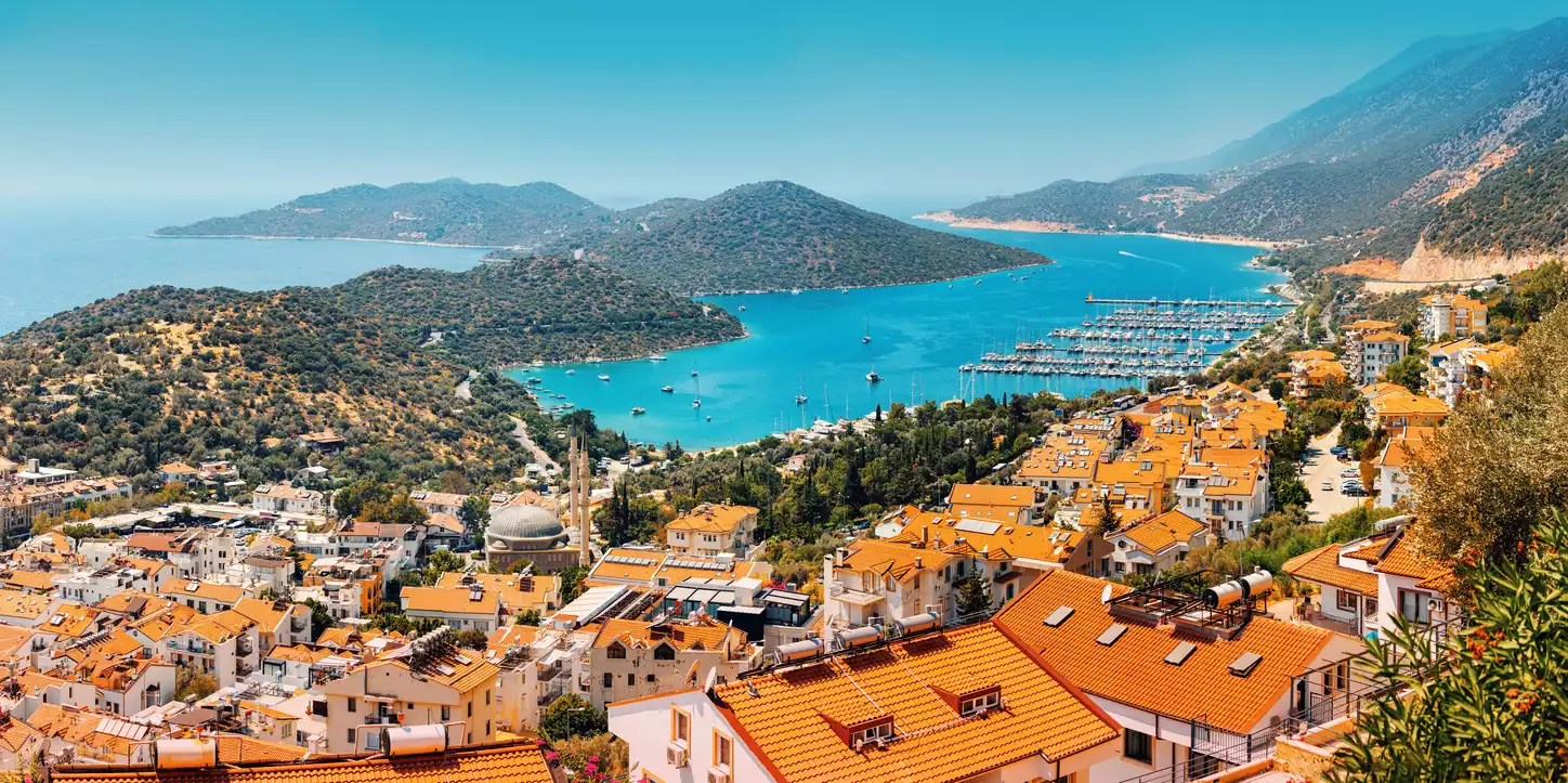 Kaş'ın tepeden çekilmiş bu geniş açılı görüntüsünde, Akdeniz'in turkuaz sularıyla çevrili, kırmızı kiremitli çatılara sahip binaların yamaca yayıldığı, birçok teknenin demirli olduğu bir marina ve yemyeşil tepelerin yükseldiği canlı bir sahil kasabası panoraması görülüyor.
