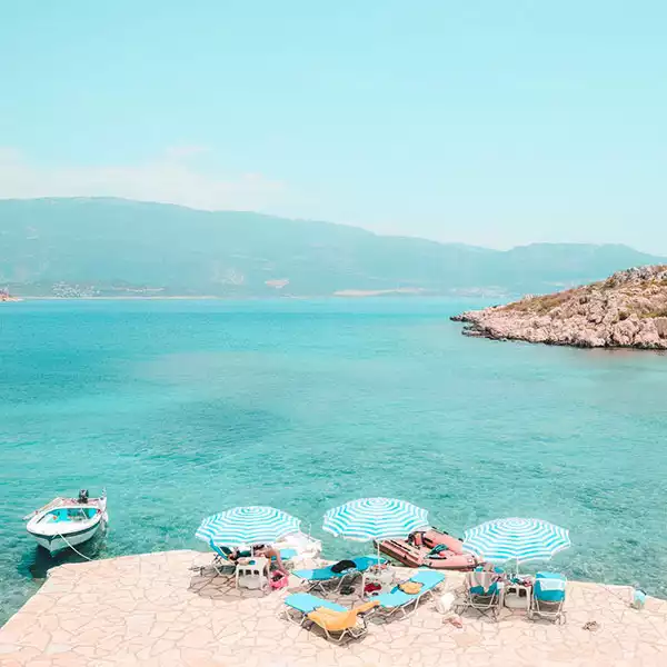 Kaş'ta, turkuaz denizin berrak sularına nazır, taş döşeli bir kıyı alanında şeritli şemsiyeler ve şezlonglarla donatılmış sakin bir koyun huzurlu manzarası, uzak dağlar ve küçük bir tekneyle çevrili doğal güzelliği gözler önüne seriyor.