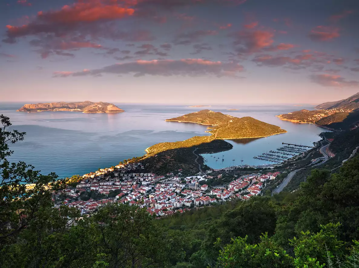 Kaş'ın gün batımında çekilmiş bu panoramik görüntüsü, şehrin Akdeniz kıyısındaki yerleşimini, kırmızı çatılı evlerini, teknelerle dolu marinasını, çevresindeki yemyeşil tepeleri ve uzaktaki adaları çarpıcı bir şekilde gözler önüne seriyor.