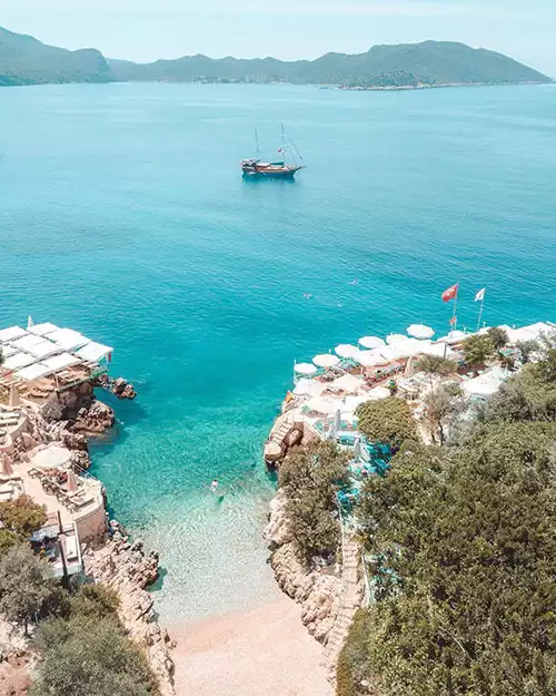 Kaş'ın berrak turkuaz sularıyla çevrili, kayalık kıyılara kurulmuş beyaz şemsiyeli ve şezlonglu şık plaj işletmelerini gösteren bu doğal koyda, sakin denizde yüzen insanlar ve uzakta yavaşça ilerleyen bir yelkenli tekne, yemyeşil tepelerin huzurlu manzarasıyla birleşerek eşsiz bir tatil atmosferi sunuyor.