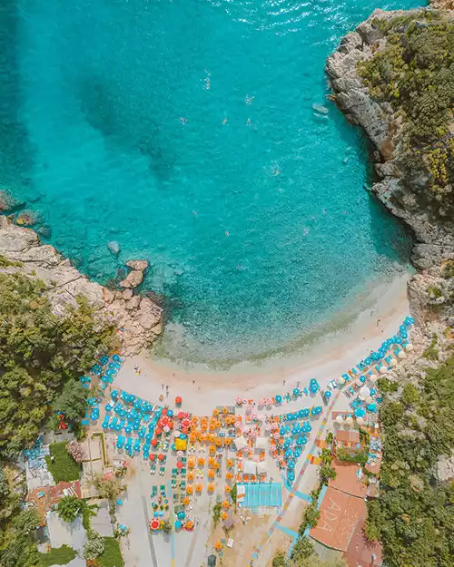 Kaş'ın berrak turkuaz sularında yüzen insanların, altın rengi kum plajında yan yana sıralanmış çok sayıda mavi, turuncu ve beyaz şemsiyeler ile şezlongların kalabalık bir görünüm sergilediği, etrafı yeşilliklerle kaplı kayalıklarla çevrili muhteşem bir koyun kuşbakışı görüntüsü.