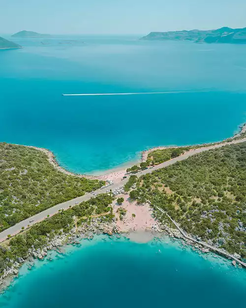 Kaş'ın turkuaz sularıyla çevrili, yemyeşil bitki örtüsüyle kaplı dar bir kara şeridinin iki ayrı koyu ayırdığı, araçların park ettiği sahil yolunun ve uzakta bir teknenin iz bıraktığı Akdeniz'in berrak denizini gösteren nefes kesici bir hava fotoğrafı.