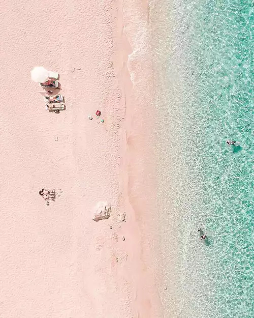 Kaş'ın pembe kumlu sahilini ve berrak turkuaz sularını kuşbakışı gösteren bu canlı fotoğrafta, plajda beyaz bir şemsiyenin altında güneşlenen ve dinlenen tatilciler ile denizin tadını çıkaran birkaç kişi dikkat çekiyor.