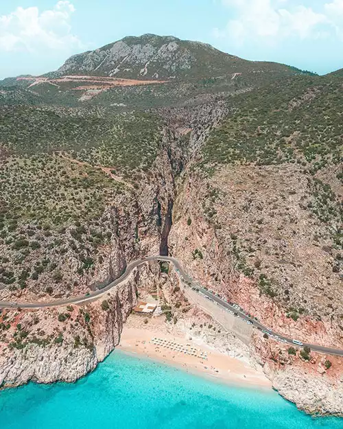 Kaş'ın simgelerinden biri olan Kaputaş Plajı'nı ve etrafındaki nefes kesici doğal güzellikleri kuşbakışı gösteren bu panoramik görüntüde, turkuaz rengi denizin, altın kumlu plajın, onu çevreleyen dik kayalıkların arasından kıvrılarak geçen yolun ve derin bir kanyonun manzarası eşsiz bir uyumla birleşiyor.