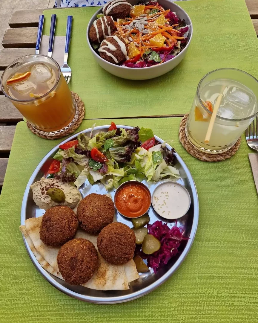 Canlı kireç yeşili bir masanın üzerinde, pide ekmeği üzerinde beş altın rengi falafel köftesi, yanında kremsi humus, taze karışık yeşil salata, canlı kırmızı lahana turşusu, çıtır salatalık turşusu ve küçük kaselerde kırmızı biber ile beyaz tahin sosu içeren bir metal tabakla dolu, beyaz sos gezdirilmiş falafeller, rendelenmiş havuç, sulu portakal dilimleri ve ay çekirdeği içeren büyük bir salata kasesi ve iki bardak ferahlatıcı buzlu içecekle donatılmış lezzetli bir sofra düzeni görülüyor.