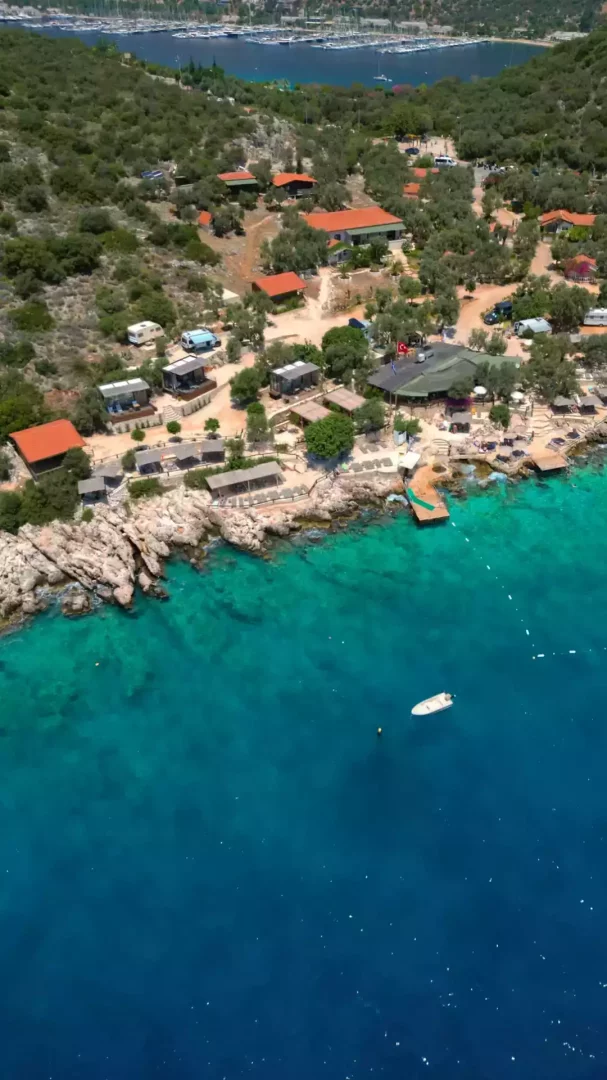 Yemyeşil tepeler ve turkuaz renkli denizin buluştuğu Kaş'ta, kayalık kıyı boyunca sıralanmış bungalovlar, karavanlar ve ahşap iskelelerin bulunduğu, dinlendirici bir Akdeniz atmosferi sunan kuşbakışı kamping alanı manzarası.