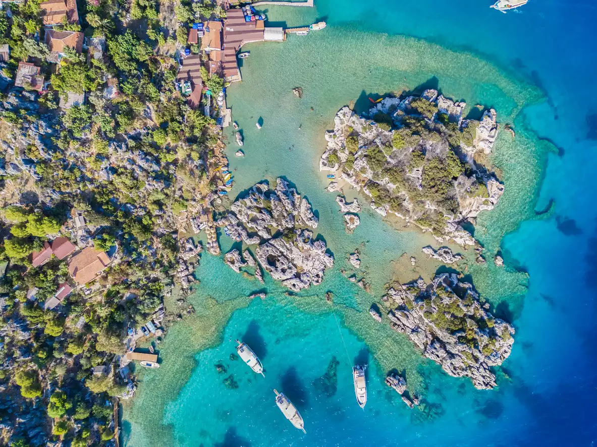 Kekova'nın kristal berraklığındaki turkuaz sularında yüzen teknelerin ve yeşil bitki örtüsüyle kaplı kayalık adacıkların kuşbakışı görüntüsü, kıyı şeridindeki geleneksel evler ve iskelelerle Akdeniz'in huzurlu atmosferini yansıtıyor.