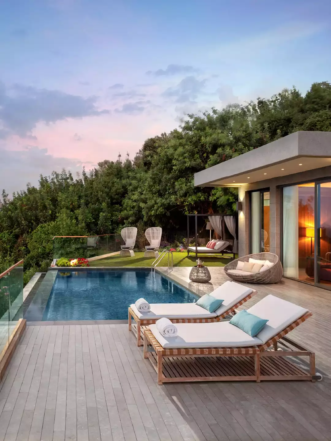 Gün batımının büyüleyici pembe ve mavi tonlarının yansıdığı gökyüzünün altında, yemyeşil ağaçlarla çevrili modern bir villa terasında, üzerinde havlular ve turkuaz yastıklar bulunan ahşap şezlongların ve parıldayan sonsuzluk havuzunun sunduğu lüks ve huzurlu bir tatil kaçamağı manzarası.
