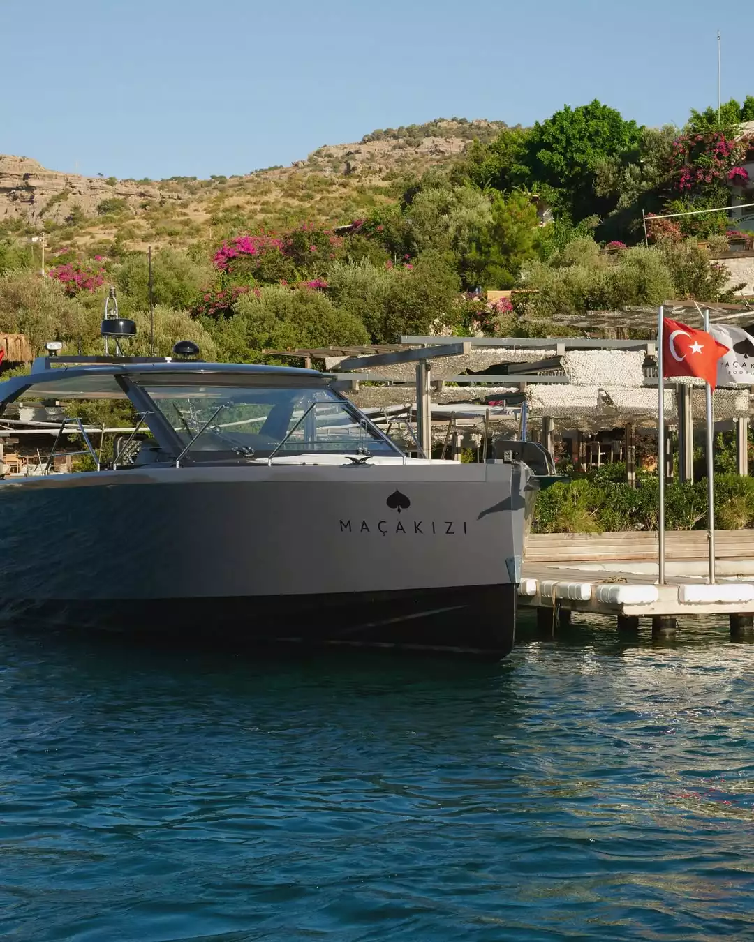 Bodrum'un göz alıcı turkuaz sularında demirli, üzerinde Maçakızı yazan modern tasarımlı gri bir yat, arkasında Türk bayrağı, pembe çiçeklerle bezeli yemyeşil yamaçlar ve tesisin gölgelikli iskele alanı ile güneşli bir günün keyfini sunuyor.