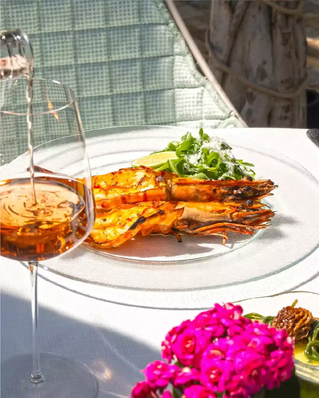 Parlak güneş altında, beyaz örtülü bir masada şeffaf tabakta duran ızgara karidesler, roka salatası, rendelenmiş peynir ve limon dilimi görünüyor; sol önde kadehe amber renkli şarap dökülürken, sağ altta canlı pembe çiçekler masaya renk katıyor.