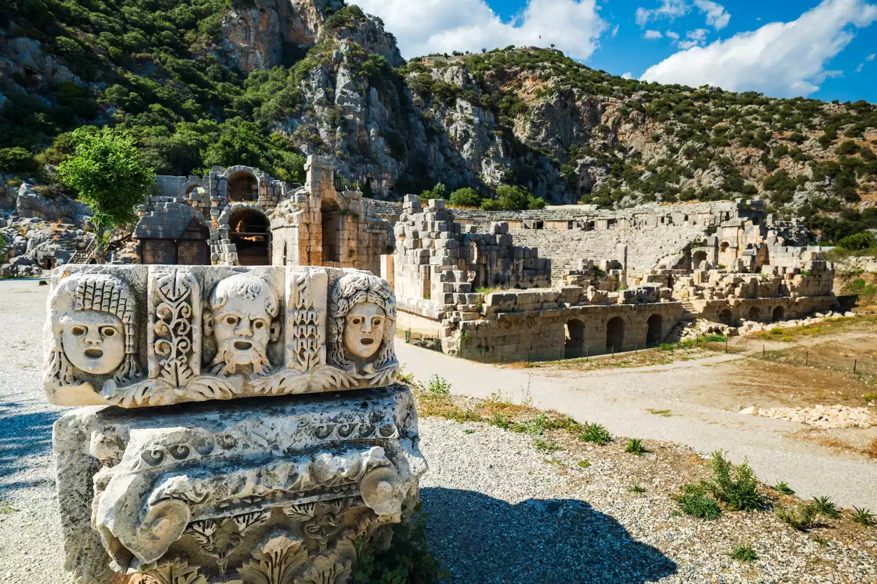 Myra Antik Kenti'nin kalıntıları arasında, ön planda insan yüzü motifleriyle işlenmiş antik bir taş blok dururken, arka planda yeşil bitki örtüsüyle kaplı kayalık dağların eteklerinde yer alan antik tiyatronun kemerli yapıları ve sıraları uzanıyor.