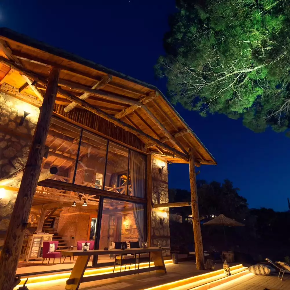 Gece ışıklarıyla aydınlatılmış, ahşap kirişli ve taş duvarlı modern bir doğa evinin geniş cam cephesi, dışarıdaki ışıklı ahşap terasıyla birleşerek huzurlu bir konaklama ortamı sunuyor, sağ üst köşede ise ışıklandırılmış büyük yeşil bir ağaç görünüyor.