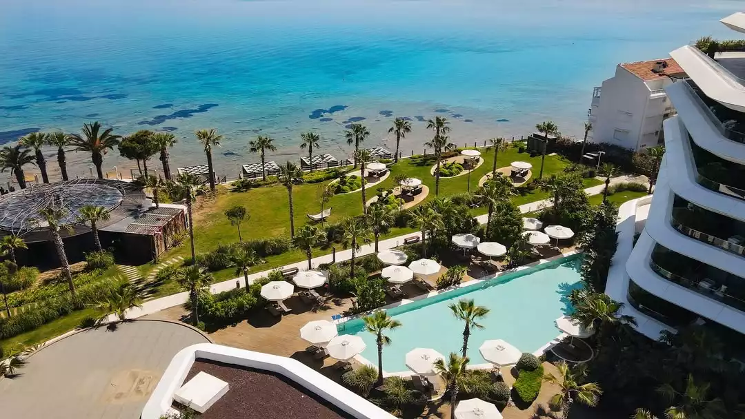 Havadan çekilmiş bu nefes kesici panoramik görüntüde, masmavi turkuaz denizin kıyısında uzanan yemyeşil bir tatil köyü, sıralı palmiye ağaçları, bembeyaz şemsiyelerle dolu şezlonglar, berrak bir yüzme havuzu ve kavisli modern mimariye sahip lüks bir otel binası yer alıyor.