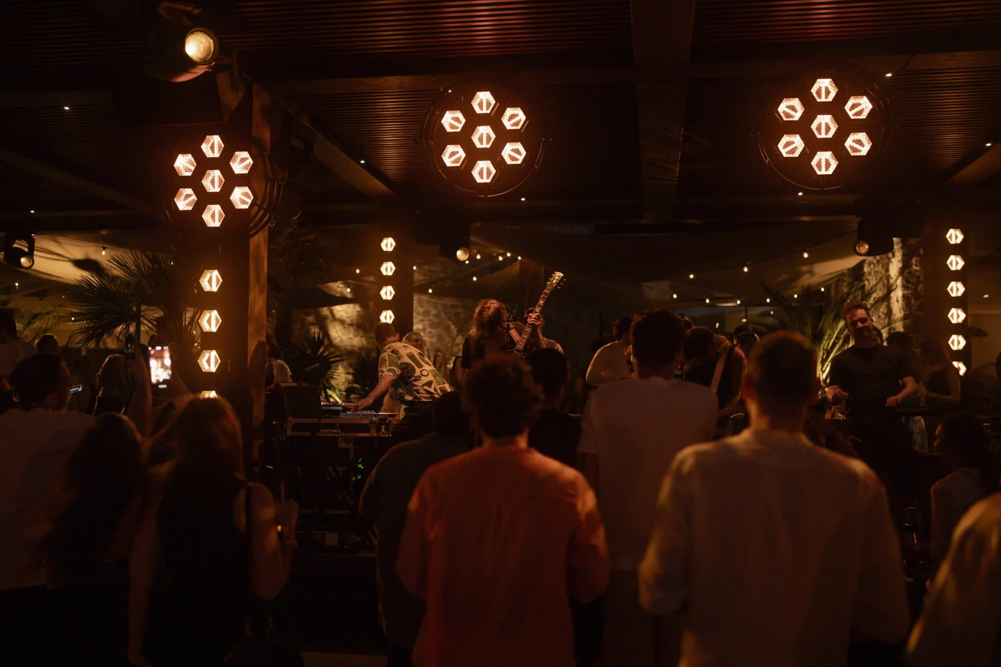 Scorpios Bodrum sahnesinde akustik gitar çalan müzisyenler ve DJ setup başında bir kişi, etraflarındaki kalabalık ile birlikte, mekana yayılan sıcak altıgen ışıkların altında dinamik bir canlı müzik performansı sergiliyor.