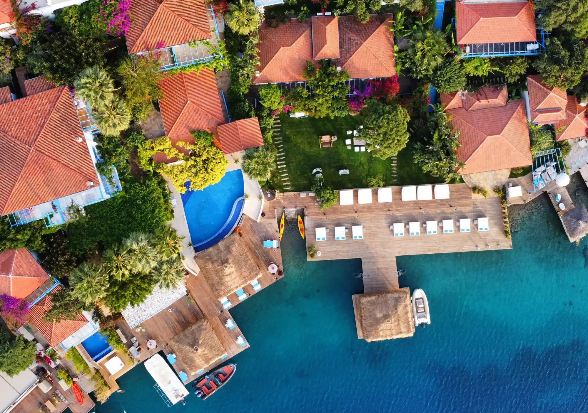 Sabrina'nın Evi'ni kuşbakışı gösteren bu etkileyici fotoğrafta, kırmızı kiremitli çatılı konaklama birimleri, tropikal bitki örtüsüyle dolu yemyeşil bahçeler, masmavi bir yüzme havuzu ve üzerinde şezlonglar, hasır çatılı cabanalar ile park etmiş tekneler bulunan turkuaz denize uzanan uzun ahşap iskeleler göz alıcı bir bütünlük oluşturuyor.