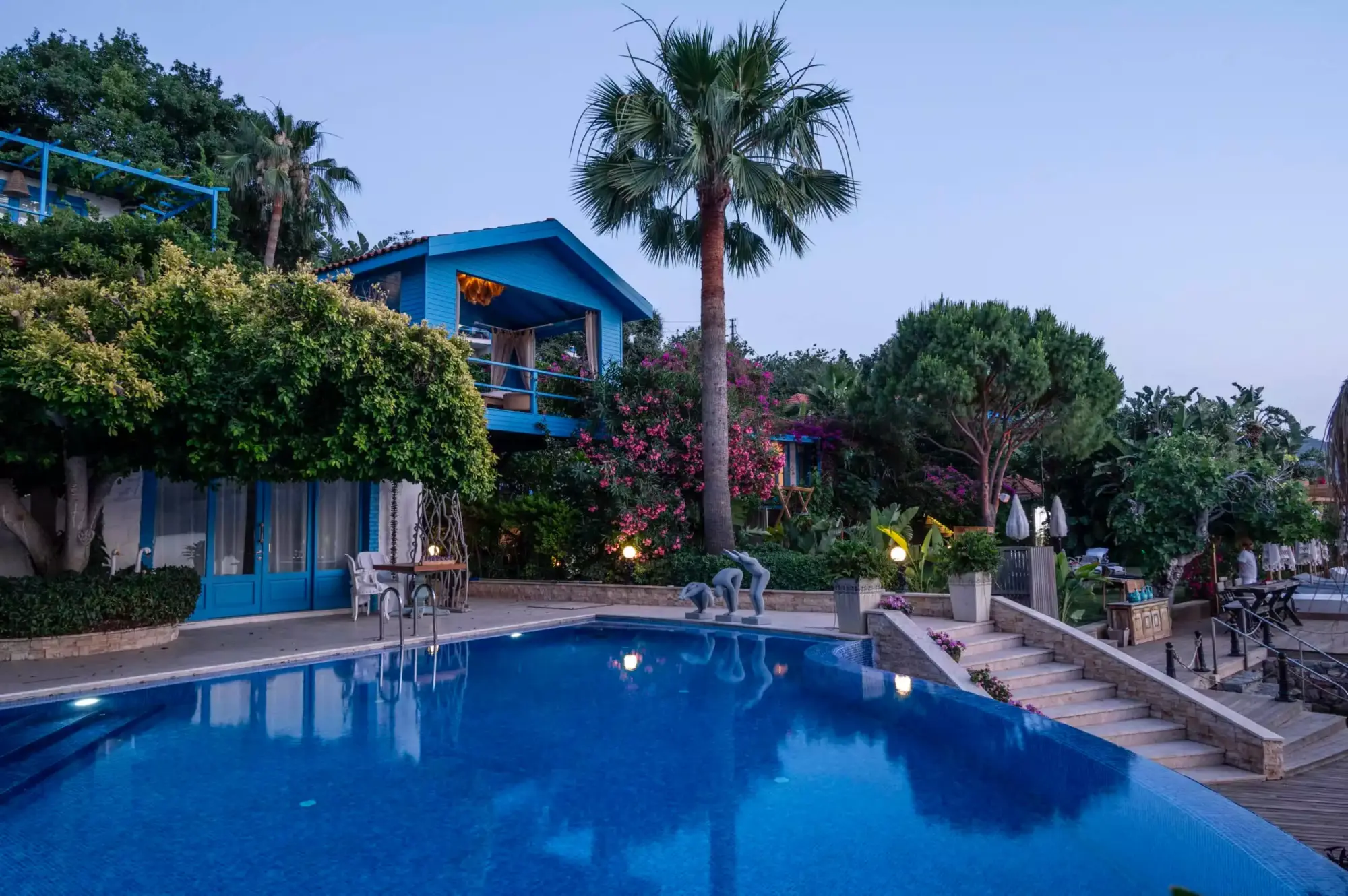Alacakaranlıkta parlayan masmavi bir yüzme havuzu, çevresinde yeşilliklerle bezenmiş, pembe çiçeklerle dolu bir bahçe ve iki katlı şirin mavi bir tatil evinin dingin atmosferini vurgulayan ışıklı heykeller ve fenerlerle çevrili Akdeniz havası sunuyor.