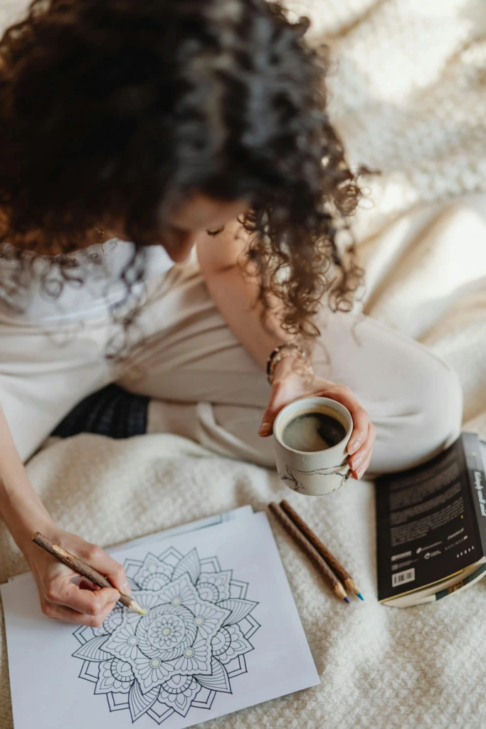 Bir kadın, battaniyesine sarınmış şekilde mandala boyarken bir yandan kahve içiyor; sanatla mindfulness anı.