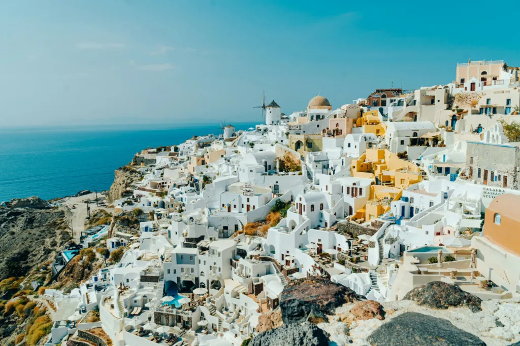 Çocuklu Aileler İçin Yunan Adaları Rehberi: Santorini'nin yamaca dizilmiş beyaz ve pastel evleri ile ikonik yel değirmeni masmavi Ege Denizi'ni kucaklıyor. Uçurumdaki bu büyüleyici yerleşim, ailelerin dar sokakları güvenle keşfederek anılar biriktireceği huzurlu atmosferi yansıtıyor.