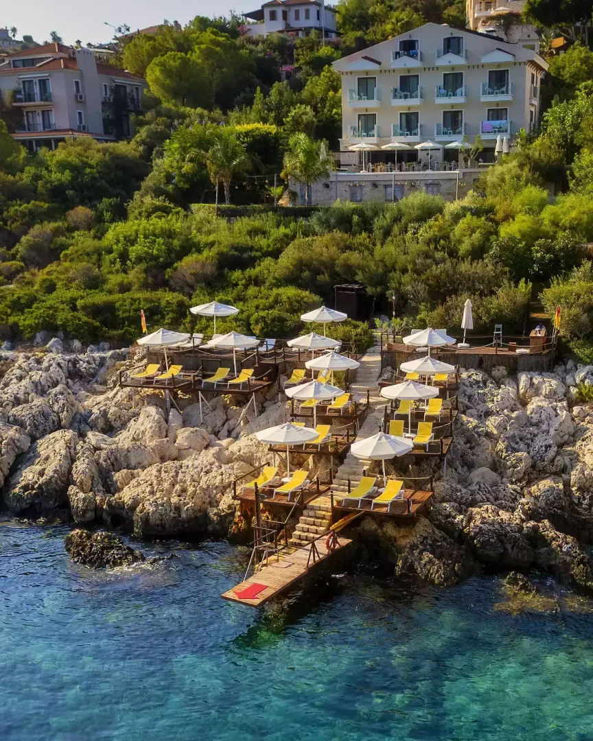 Yeşil ağaçlarla kaplı bir tepenin yamacına konumlanmış Suna-Sun Hotel'in genel görünümünü gösteren bu geniş açılı çekimde, otelin altındaki kayalık kıyı şeridinde, berrak turkuaz denize doğru kademeli olarak inen, sarı şezlonglar ve beyaz şemsiyelerle donatılmış çok sayıda ahşap güneşlenme terası ve suya uzanan küçük bir iskele dikkat çekiyor.
