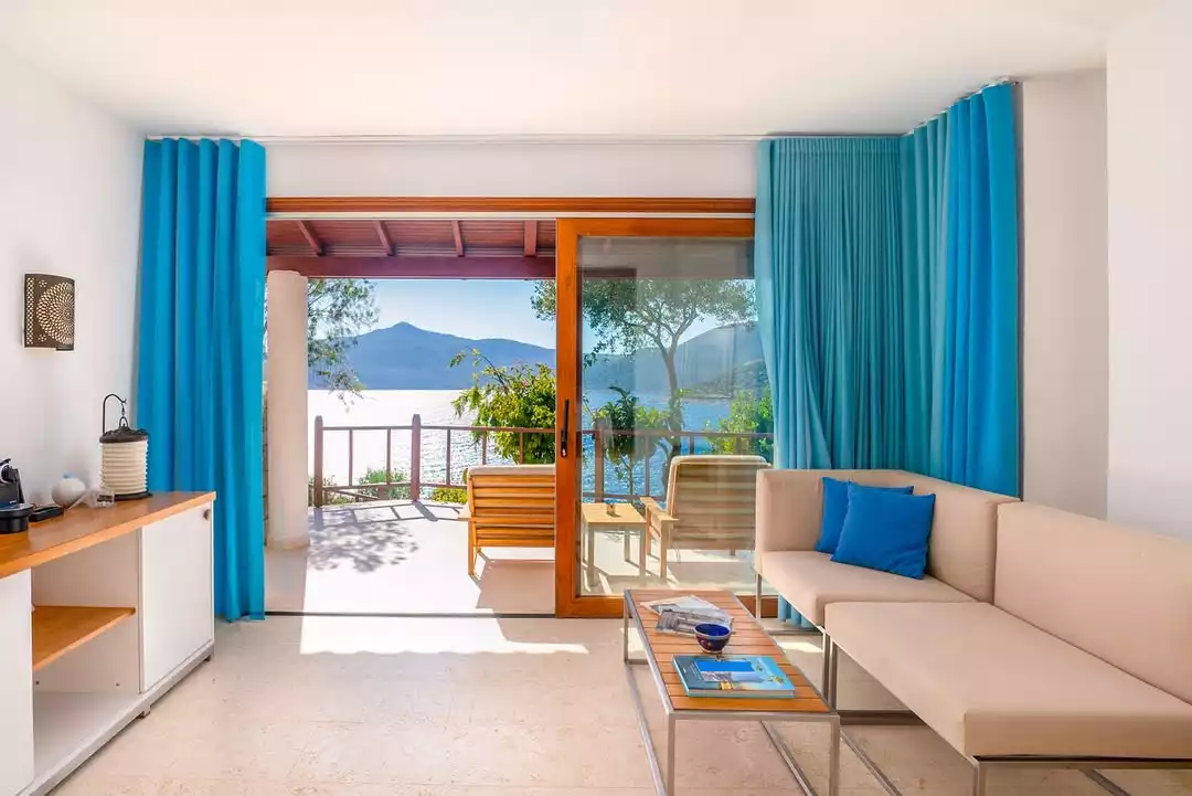 Villa Mahal'in denize açılan geniş balkonlu, modern ve aydınlık oturma odasında, turkuaz perdelerle çevrili sürgülü cam kapıdan Akdeniz'in berrak suları ve yeşil dağ manzarası eşliğinde ferah bir ortam sunuluyor; iç kısımda bej renkli rahat bir koltuk ve dekoratif bir sehpa göze çarpıyor.