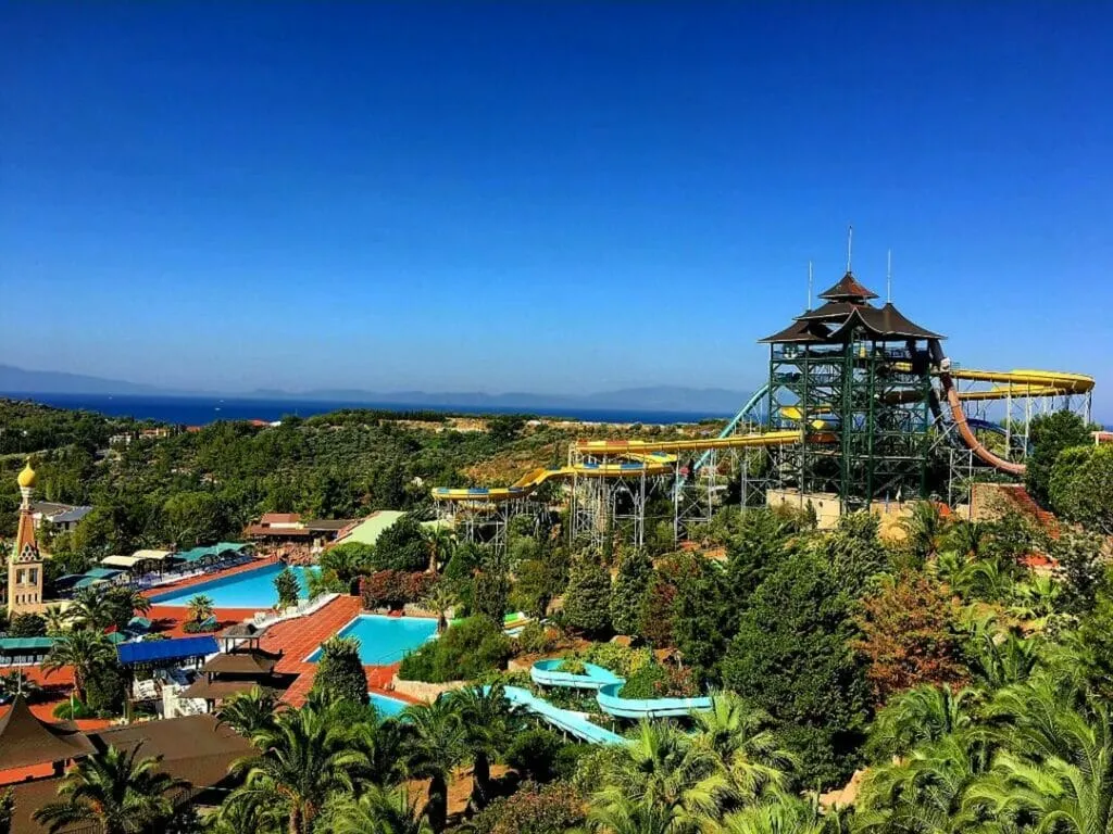 Kuşadası yakınlarındaki Adaland su parkının genel görünümünde, çok katlı kaydırak kulesi, çeşitli su kaydırakları, mavi yüzme havuzları ve palmiye ağaçlarıyla dolu yemyeşil tesisler, masmavi Ege Denizi manzarasıyla birleşerek keyifli bir eğlence ortamını sergiliyor.