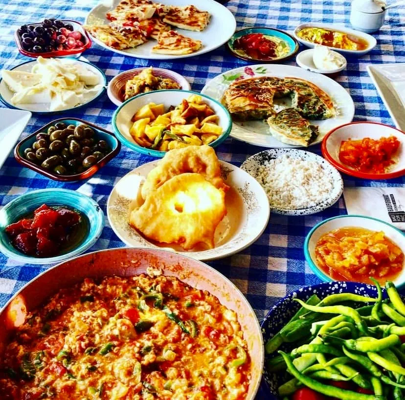 Asmalı Çardak'ta sunulan zengin bir Türk kahvaltısı masası, menemen, pişi, zeytin, peynir, börek, reçeller ve taze yeşil biberler gibi birçok lezzetli çeşidin, mavi-beyaz kareli örtü üzerinde iştah açıcı bir şekilde düzenlenmiş yakın çekim bir görüntüsüdür.