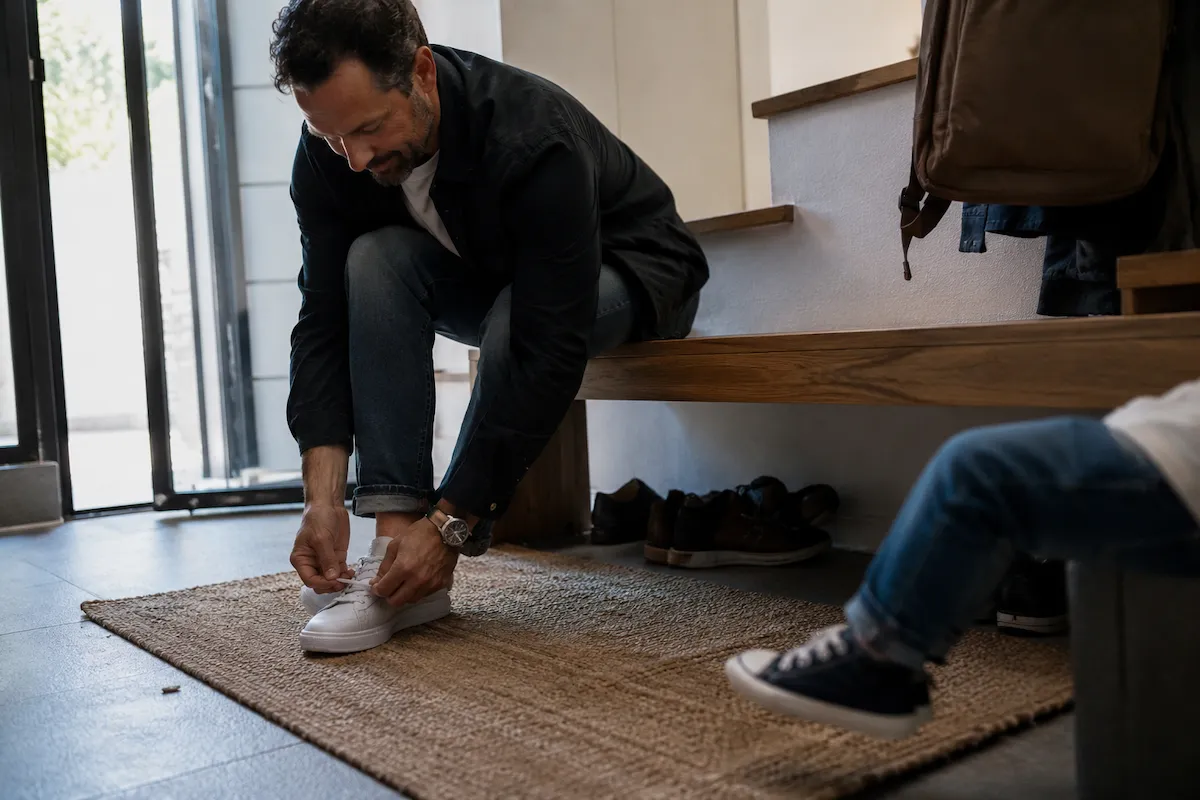Bir baba, evinin girişindeki ahşap bankta oturmuş, gündelik baba stili tamamlayan beyaz spor ayakkabılarının bağcıklarını dikkatle bağlıyor; arka planda ise küçük bir çocuğun ayakları ve çeşitli ayakkabı seçeneklerini ima eden diğer ayakkabılar da seçiliyor.