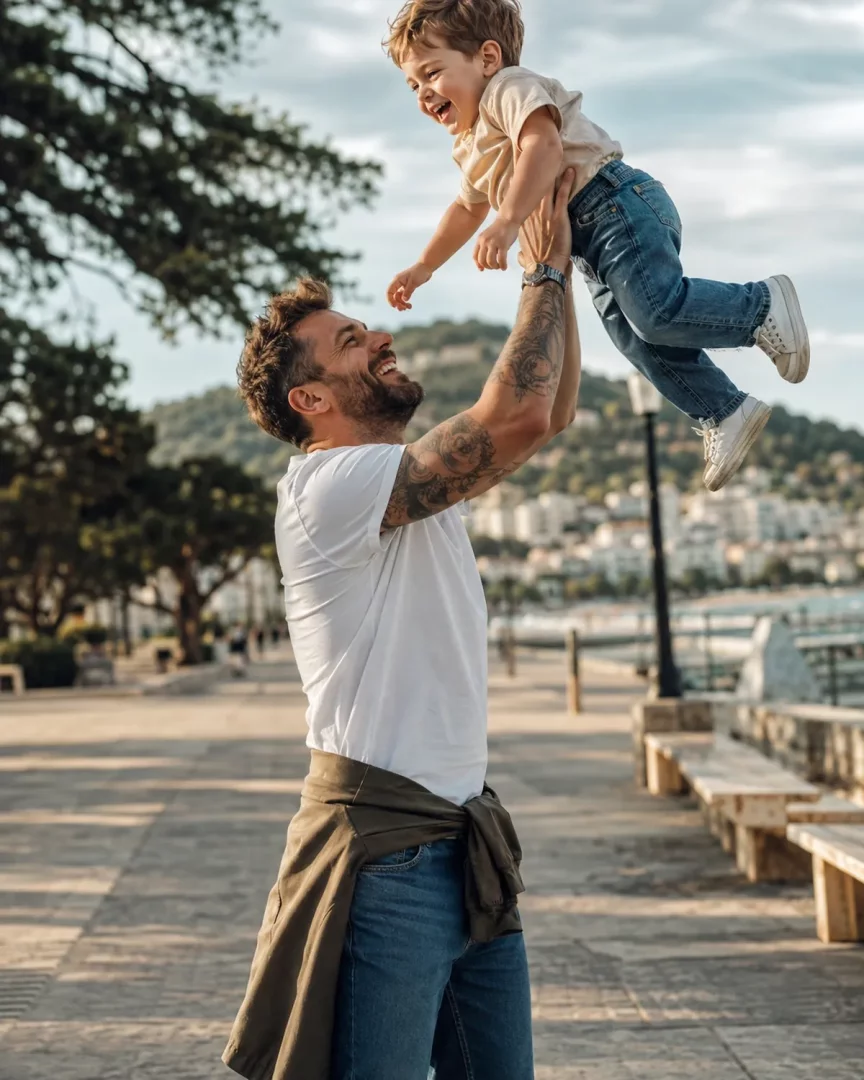 Gülümseyen ve dövmeli bir baba, modern günlük stilini yansıtan baba stili beyaz tişörtü ve jean pantolonuyla, kollarını havaya kaldırdığı kahkahalar atan küçük çocuğunu neşeyle izlerken sahilde keyifli bir baba-çocuk anı yaşıyorlar.