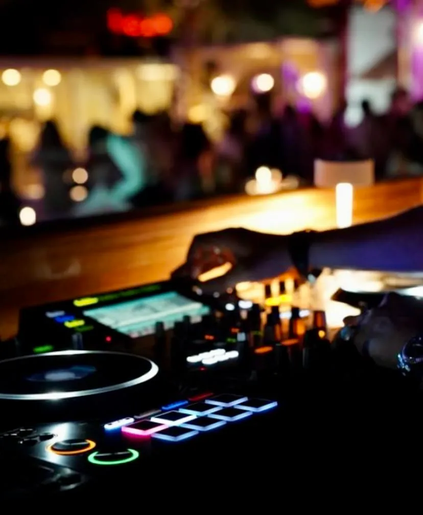 Karanlık bir ortamda, DJ performansı sırasında elleri ışıklı tuşları ve pikapı kullanan bir DJ'in mikseri ve kontrol paneli ön planda belirginleşirken, arka planda mekanın sıcak ambiyansını yansıtan bulanık sarı ve mor ışıklarla dolu bir kalabalık görülüyor.